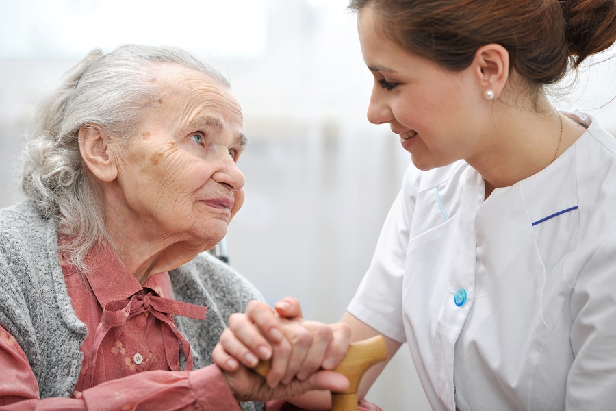 Nurse smiling at an elderly woman, gently holding her hands for support.