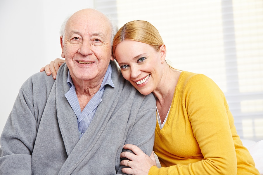 Smiling elderly man in a gray robe sits with a younger woman in a yellow sweater leaning close, arm around him.